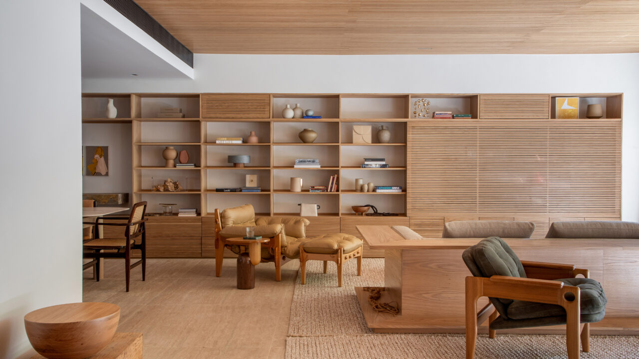 Materiais naturais e tons claros protagonizam projeto do PKB Arquitetura. Na foto, sala com estante de marcenaria, poltrona e sofá.