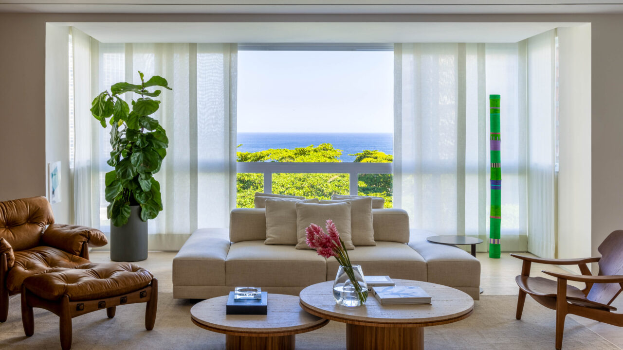 Mar de São Conrado é enquadrado nas janelas deste projeto de LessaZanetta. Na foto, sala de estar com estante de madeira, sofá e poltronas.